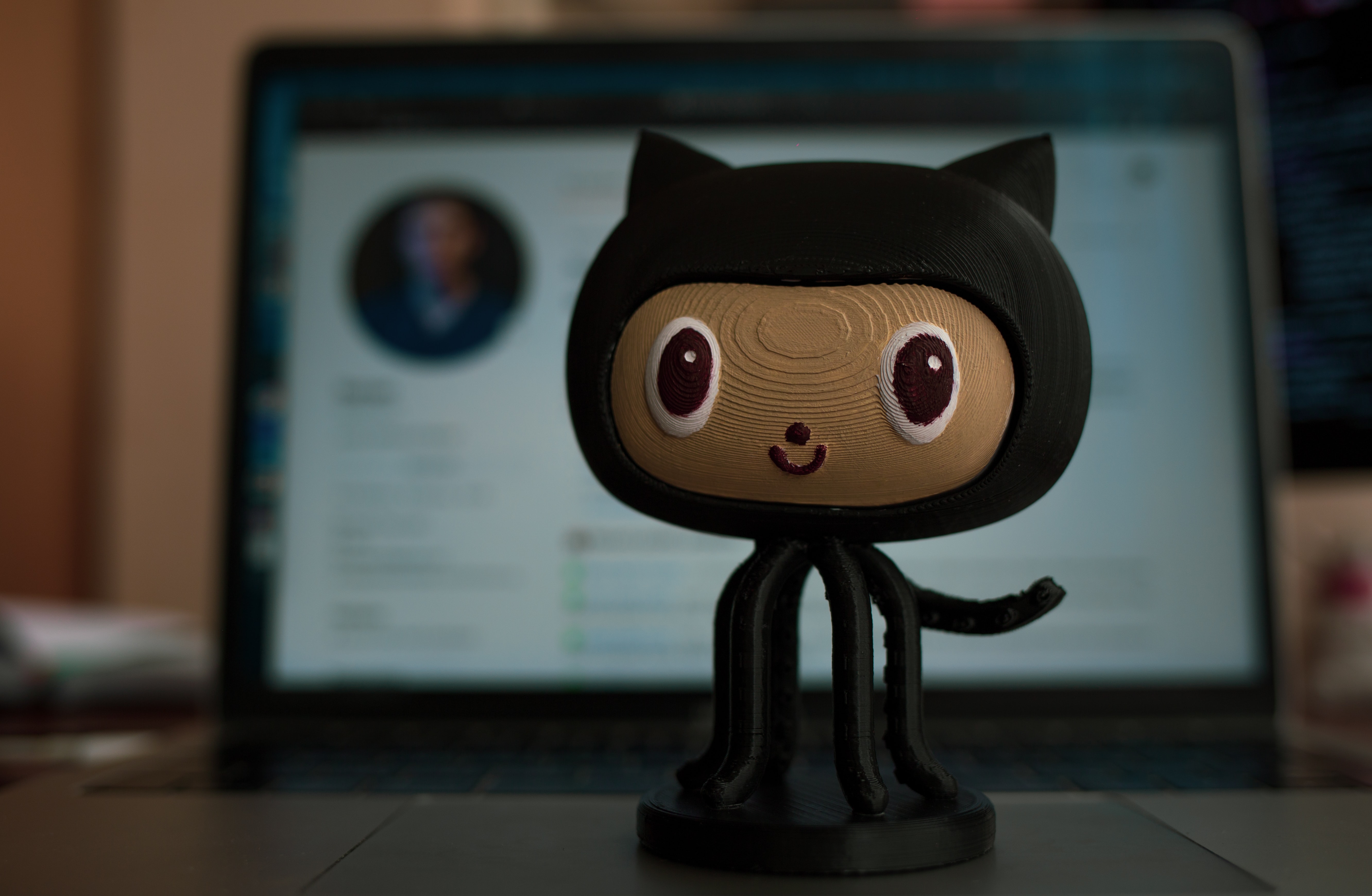 Uma estatueta de um oktokat no centro, ao fundo um laptop com a página principal do GitHub aberta